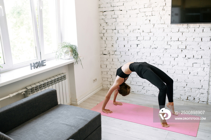 Young fit girl stands in a bridge while a home workout. She is doing her exercises at home on a fitness mat, stretching, healthy habbits