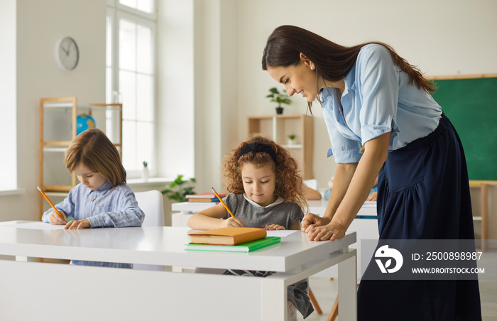 Education for the youngest. Friendly Caucasian teacher helps the junior students to complete the school task correctly. Smiling teacher in class walks between desks and teaches young students to write