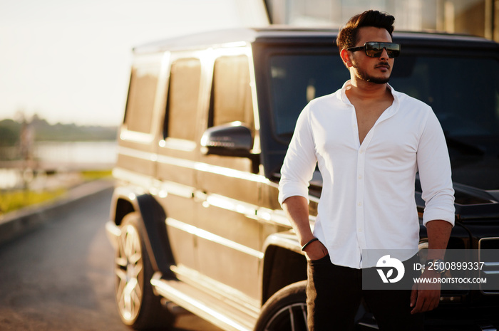 Solid asian man in white shirt and sunglasses posed near black mafia suv car.