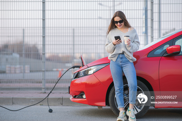 Electric car charging in street. Ecological Car Connected and Charging Batteries. Girl Use Coffee Drink While Using SmartPhone and Waiting Power Supply Connect to Electric Vehicles for Charging