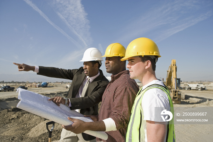 Side view of a surveyor and construction workers in hard hats with plans on site