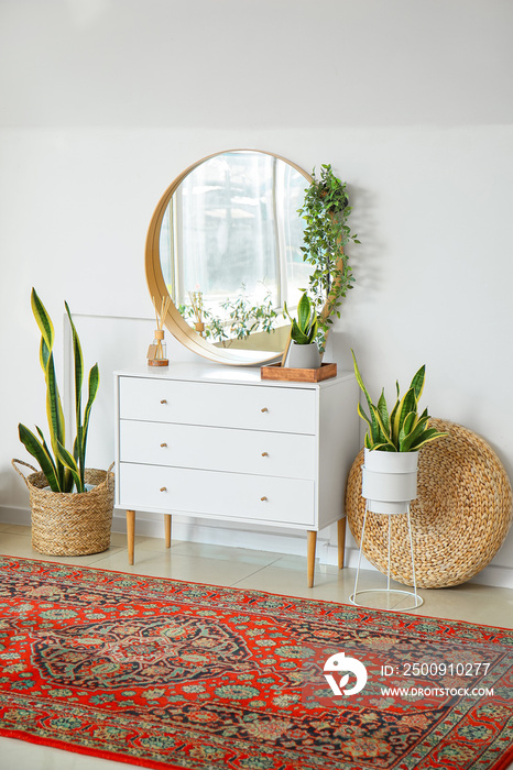 Chest of drawers with houseplants, mirror and pouf near light wall