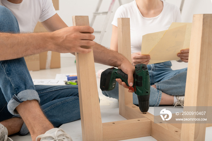 Close-up of unrecognizable man sitting on floor and using screwdriver while assembling table with wi