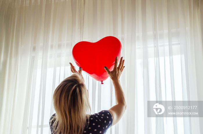 Young woman holding red heart shaped balloon against the window at home. Celebrating valentines day,