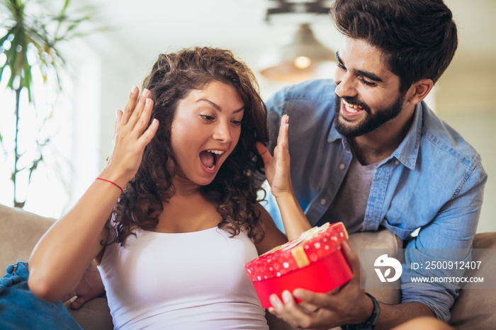 Beautiful young couple is celebrating at home. Handsome man is giving his girlfriend a gift box