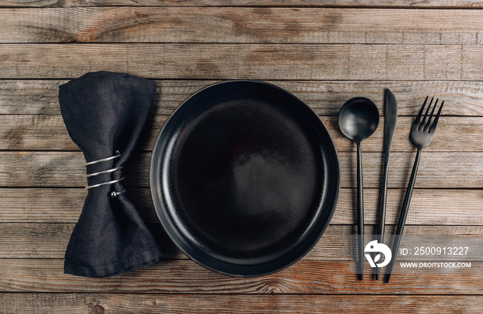 Black plate, cutlery and napkin on rustic wooden background. Table setting.