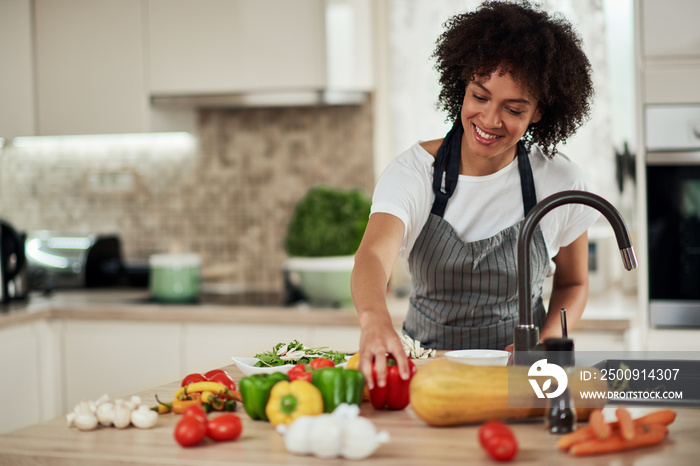 Beautiful mixed race houswife reaching for red pepper while standing in kitchen. Dinner preparation.