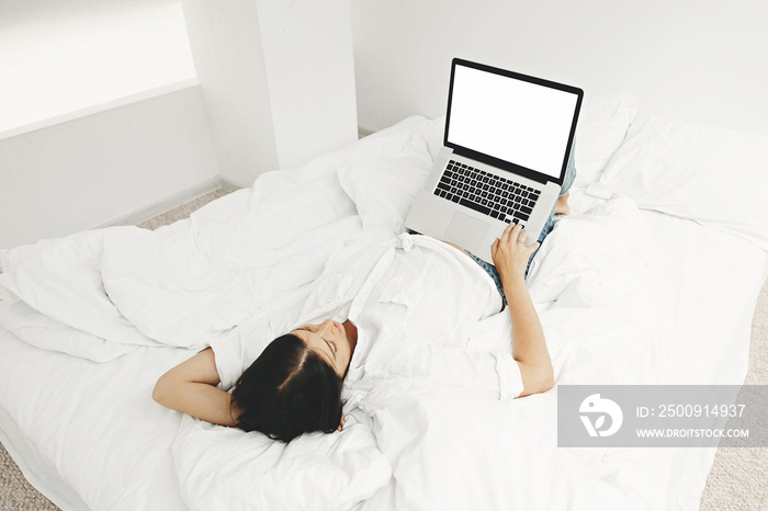 Happy stylish girl lying on bed with laptop with blank screen in white modern room. Young woman look