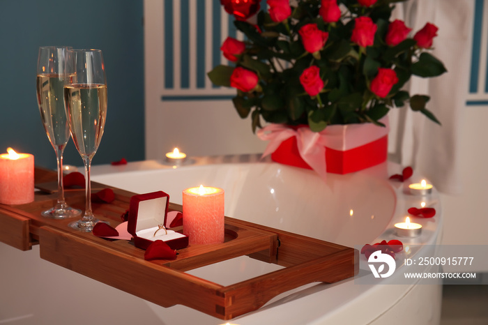 Tray with engagement ring, glasses of champagne and burning candles on bathtub in room decorated for