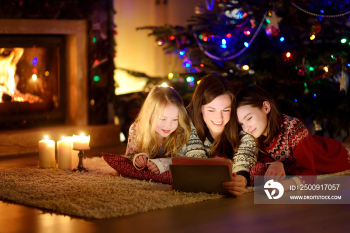 Mother and her two cute young daughters using a tablet pc at home by a fireplace in warm and cozy li