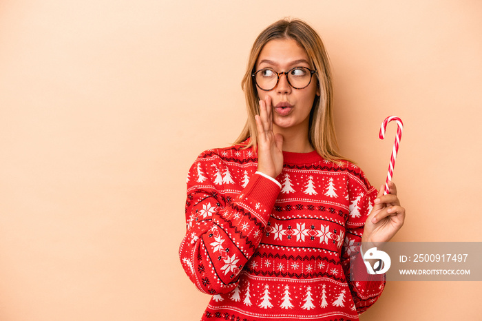 Young caucasian woman holding a christmas stick isolated on beige background is saying a secret hot 