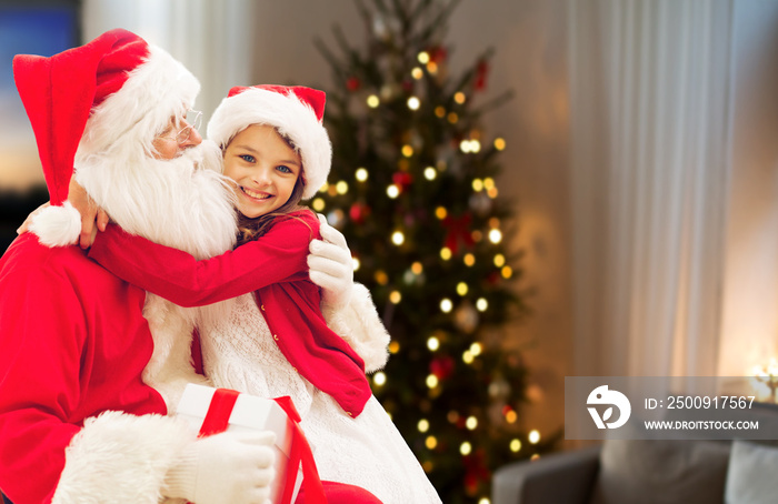 holidays, childhood and people concept - smiling girl hugging santa claus with gift over christmas t