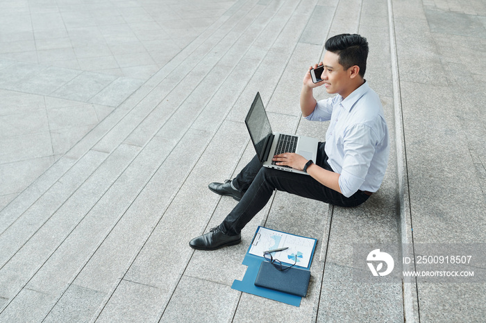 Cheerful young Vietnamese businessman sitting on steps, working on laptop and calling on phone