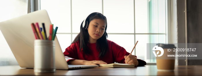 Female student wears headphones sitting at table to learning and note, using laptop for homework dur