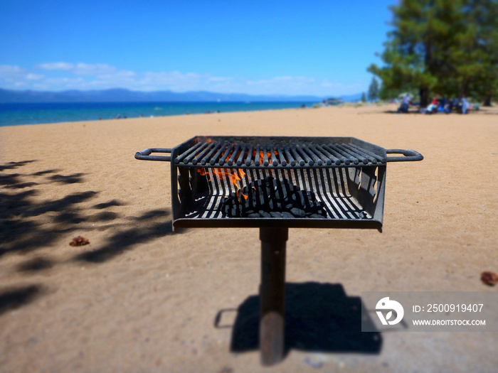 Charcoal grill with flames on a beach in Lake Tahoe with the lake in the background on a sunny day