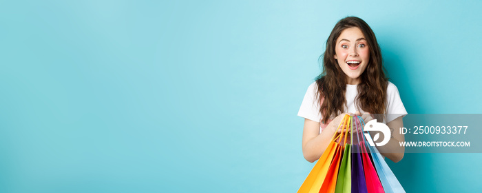 Portrait of excited girl shopper, holding shopping bags, buying in stores and smiling amazed, enjoy discounts, standing over blue background