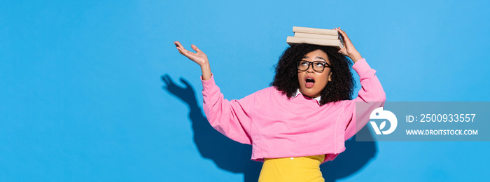 amazed african american woman with books on head pointing with hand on blue, banner