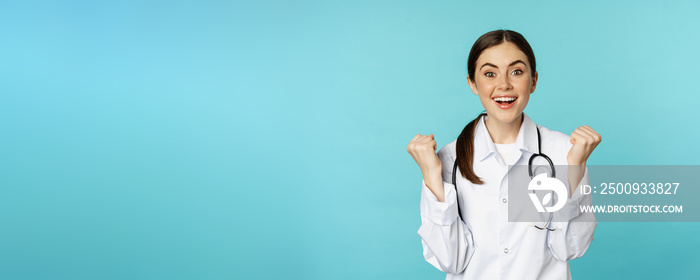 Happy and excited young female doctor, physician celebrating, achieve goal and smiling pleased, winning, standing in white coat against blue background