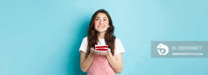 Holidays and celebration. Dreamy birthday girl making wish and looking up hopeful, holding bday cake and smiling, standing over blue background