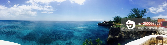 Panoramic view from Negril, Jamaica (Caribbean island): turquoise blue ocean, sky with white clouds, palm trees, stone wall and red house create a beautiful summer vacation