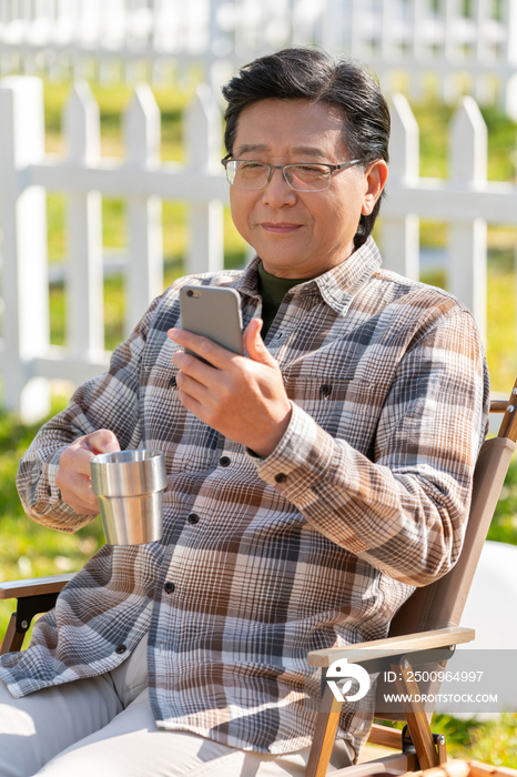老年男人坐在露营院子里看着手机视频喝咖啡