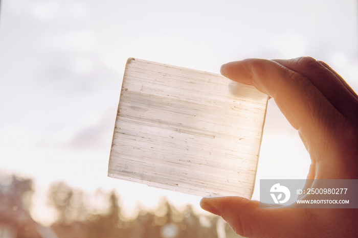 Person holding natural mineral stone Selenite plate against sun and blue sky, stone has healing and cleansing properties. Also used for recharging other mineral crystal stones.