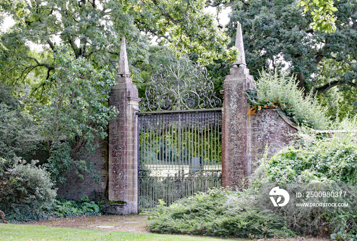 Old wrought iron gates mounted between two tall slim brick gate posts in an English country garden.