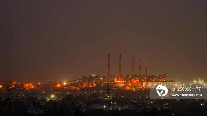 Night urban landscape with an industrial complex with its metal structures and chimneys with reflection of lamps with yellow lights of the city.