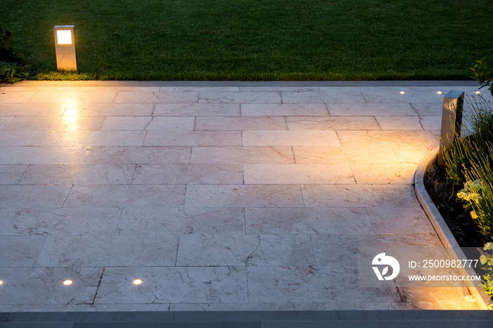 The pavement of marble tiles and a stone border area in the evening illuminated by ground lanterns in a metal housing shining a warm light around the lawn.