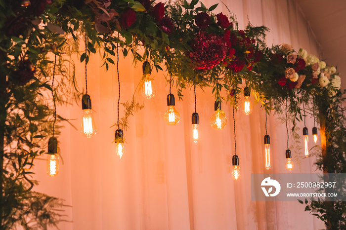 Wedding reception decoration with different electric edison lamps and fresh red and bordo flowers, rustic style