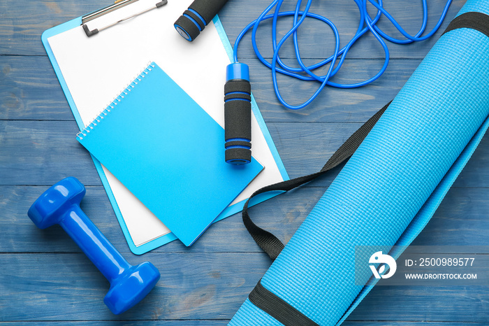 Dumbbell with notebook, clipboard, yoga mat and skipping rope on wooden background