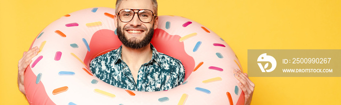 Smiling bearded guy in glasses with donut swim ring isolated on yellow, panoramic orientation