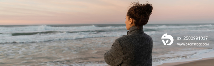 Curly woman in coat looking away while standing on beach near sea in Spain, banner.