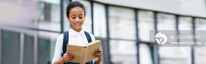 smiling african american schoolgirl reading book outdoors, panoramic shot