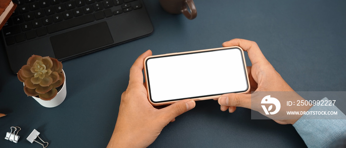 Hands of businessman holding horizontal smart phone with empty screen on dark blue leather.