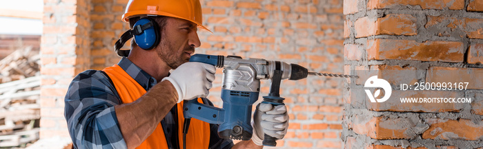 panoramic shot of handsome bearded man using hammer drill near brick wall