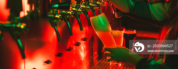 Young woman barman pouring a cold lager beer from tap to glass in neon light.