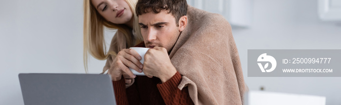 blonde woman holding blanket near boyfriend with cup working remotely from home, banner.