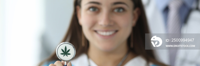 Close-up of female hand holding stethoscope with cannabis symbol. Cheerful smiling nurse in medical uniform in clinic. Healthcare and medical marijuana concept