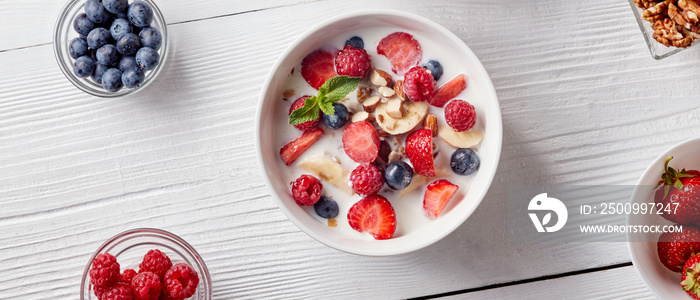 Cereal muesli, banana, blueberries, raspberries, almonds and walnuts with milk in white ceramic bowl on a white table. Top view.