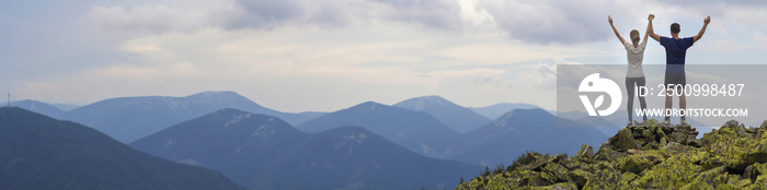 Panorama of young couple, athletic boy and slim girl standing with raised arms on rocky mountain top enjoying breathtaking summer mountain view. Tourism, success and healthy lifestyle concept.