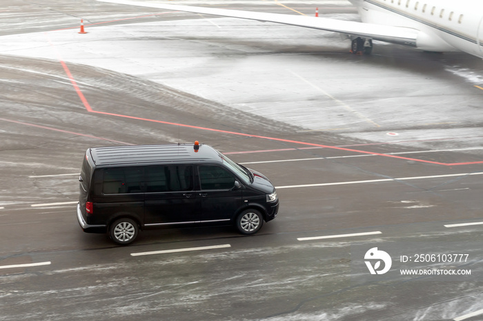 黑色贵宾服务车在机场滑行道上行驶，背景是模糊的私人飞机。商务cl