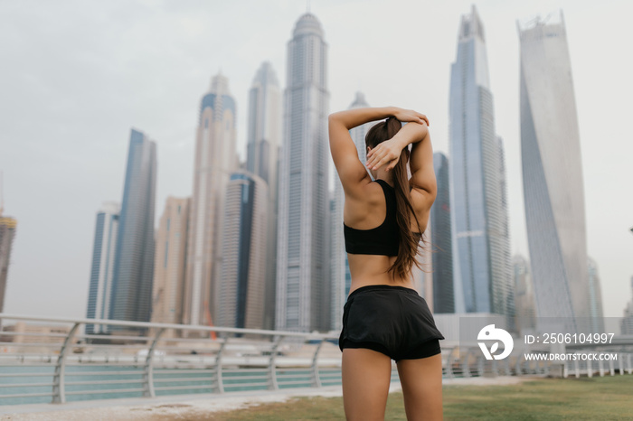 Young sporty girl wearing sportswear stretching arms near skyscrapers, preparing for working out, ou