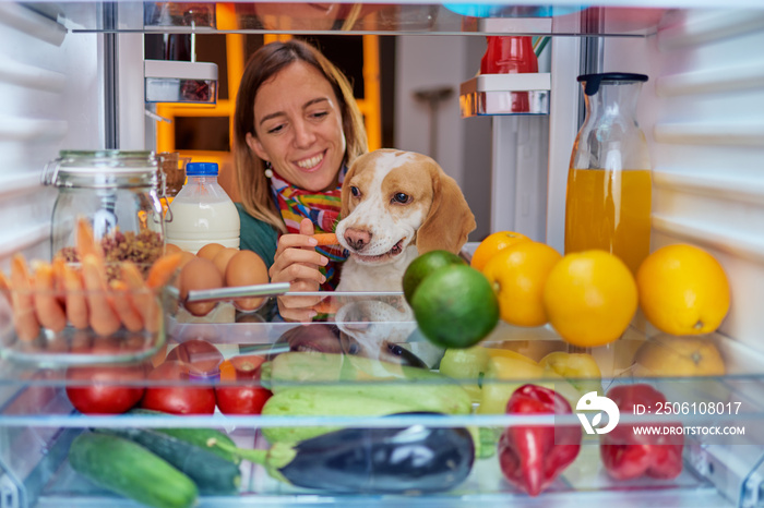 Woman feeding dog while standing in front of fridge full of groceries. Picture taken from the inside