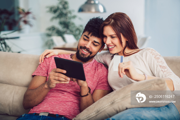 Happy mixed race couple shopping online at home, using credit card, laughing