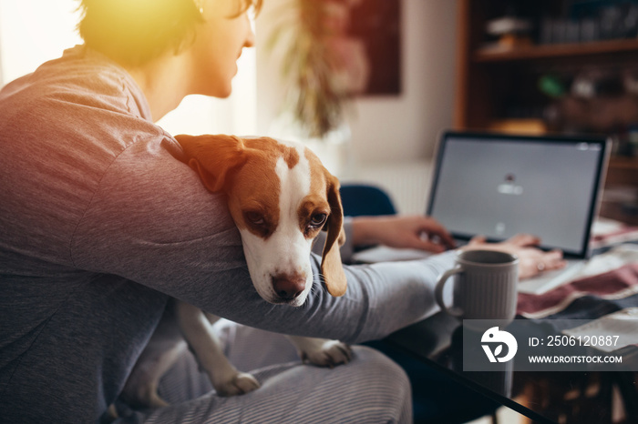 young man cuddling with his dog while drinking morning coffee and using laptop in his kitchen