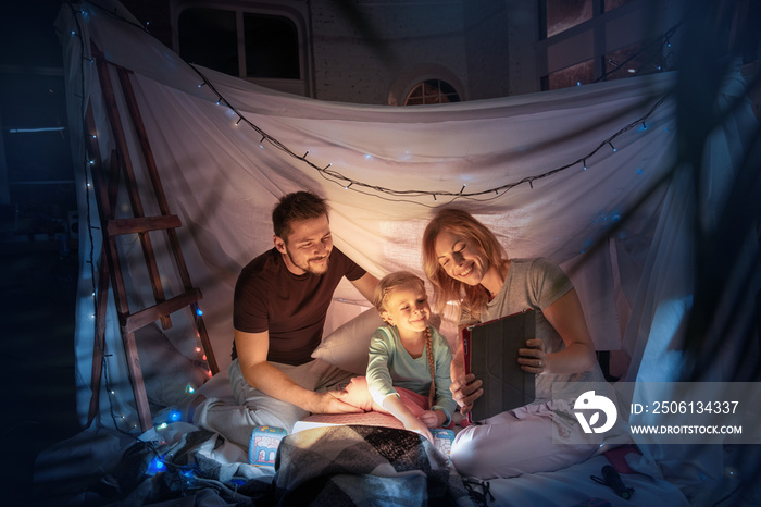 Caucasian family sitting in a teepee, having fun and playing with the flashlight in dark room with t