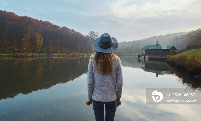 Stylish young woman in a warm sweater and hat, standing on pier by the lake in the park at autumn. B