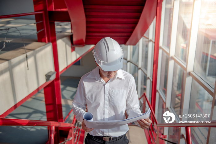 年轻的亚洲男工程师或建筑师在红色st上戴着防护安全帽拿着蓝图