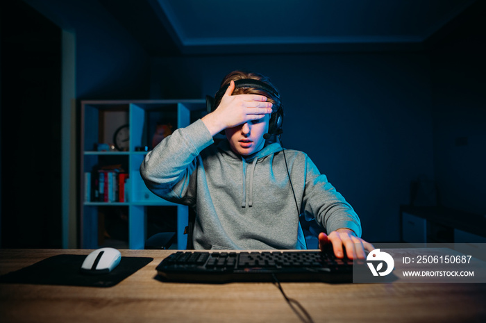 gamer loser in a headset sits at night in a room with a blue light at a table at a computer and make